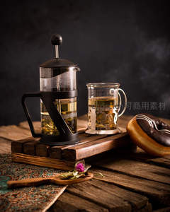 法压壶与玻璃杯盛装清亮绿茶，搭配巧克力甜甜圈与干花点缀木质托盘