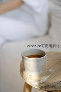 粗陶茶杯置于原木凳上，茶包悬垂，背景虚化白衣人影