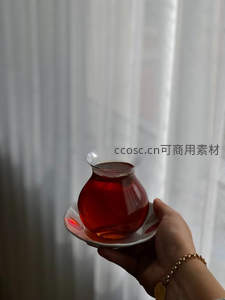 手持彩绘茶盏托起琥珀红茶，薄纱窗帘透光营造柔美私享时光