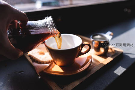 窗边阳光下，手执玻璃瓶倾倒冷萃咖啡入陶杯，配饼干与金属奶盅，光影温暖