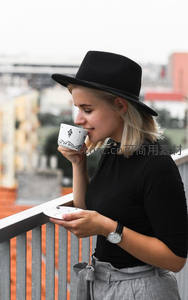 都市阳台品咖：戴黑帽女子轻啜手绘咖啡杯的惬意时光