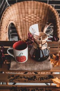 秋日氛围手冲咖啡：南瓜图案马克杯与玻璃滤壶置于藤椅木桌，落叶点缀怀旧风