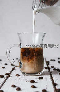 牛奶缓缓注入冰美式玻璃杯，咖啡豆散落白色木板桌面