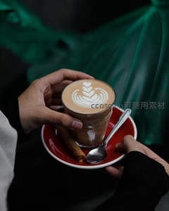 双手捧红色瓷碟咖啡杯，拉花心形叶纹，搭配肉桂棒与金属勺