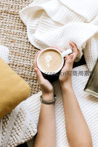 双手捧着拉花咖啡杯，毛毯与针织纹理营造温馨居家氛围