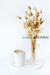 干花兔尾草插于玻璃瓶旁，搭配拉花咖啡杯，极简白调静物美学