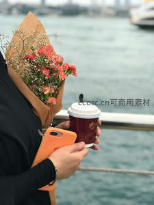 手持花束与咖啡杯临海而立，橙色手机映衬浪漫都市慢旅行