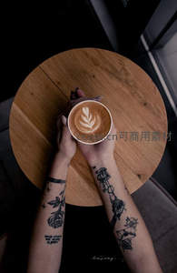 纹身双手捧着拉花咖啡杯，木质桌面与暗调背景形成对比