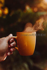 暖光下手持橙色咖啡杯，热气袅袅升腾于自然背景前
