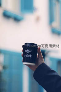 手持印有'coffee o'clock'字样的黑色纸杯，蓝白建筑背景虚化