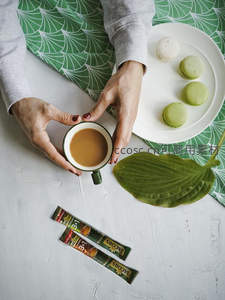 双手捧速溶咖啡杯配马卡龙，绿叶桌布与咖啡条包构成春日下午茶场景