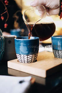手冲咖啡正注入蓝釉几何纹陶杯，木质托盘与暖光营造精品咖啡仪式感
