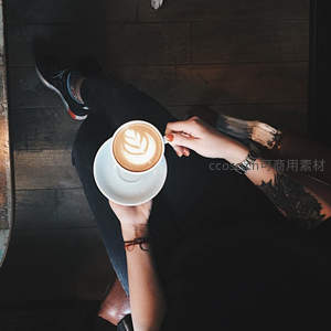俯拍视角下，纹身手臂轻握拉花咖啡杯，木质地板与休闲鞋构成慵懒咖啡时光