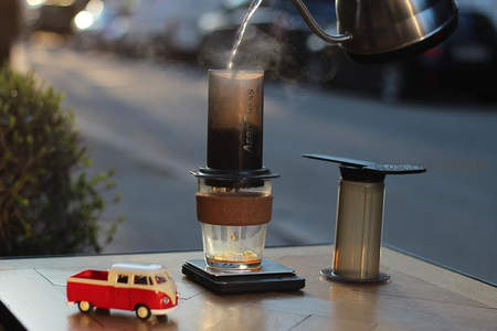 手冲咖啡制作瞬间：热水注入AeroPress滤杯，蒸汽升腾，旁置复古小车模型