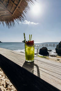 海边木桌上的绿色鸡尾酒，配热带茅草伞与碧海蓝天背景