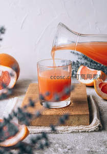玻璃壶倾倒西柚汁入杯，木板衬托切片西柚与干花点缀