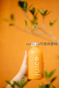 手握橙汁瓶置于暖橙背景前，绿植虚化衬托清新活力感