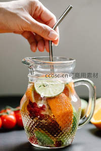 手握金属吸管搅动水果冷泡茶，玻璃壶中橙片青柠与浆果层次分明