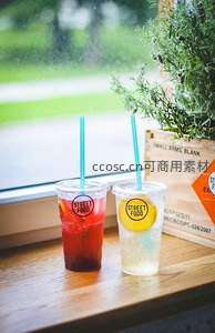 窗边木桌上两杯街食品牌冷饮，红黄双色搭配绿植背景清新自然