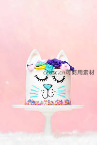 猫咪造型蛋糕带闭眼笑脸、彩色糖珠与花朵头饰，粉背景可爱风格
