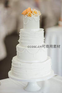 四层纯白婚礼蛋糕配褶皱糖霜与珍珠链饰，顶端橙色玫瑰优雅绽放