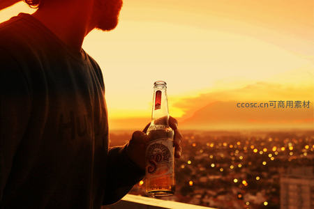 男子手持Sol啤酒眺望城市日落，剪影轮廓与万家灯火交织成惬意夜景