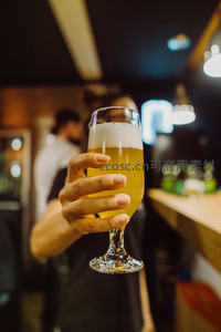 手持精酿啤酒杯特写，金黄酒液泛白沫，背景虚化吧台与人影营造温馨社交场景