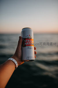 日落海边手持 Stella Artois 啤酒罐，惬意度假时刻