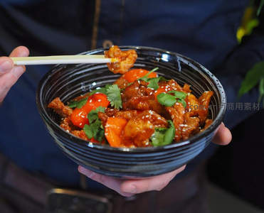 手持黑釉碗盛糖醋里脊，鲜亮酱汁裹肉块配彩椒香菜与芝麻