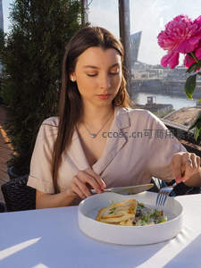 窗边女子专注品尝煎鱼沙拉，城市河景与牡丹花映衬优雅用餐时刻