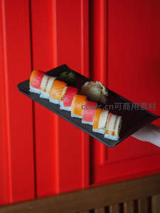 黑盘盛放三文鱼与金枪鱼寿司卷，配芥末姜片，红墙背景衬托鲜亮色泽