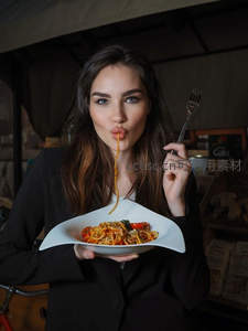 黑衣女子俏皮吸食意面，手持异形白盘，背景市集摊位增添生活烟火气