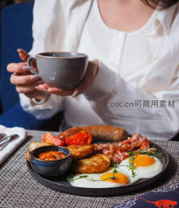 女士手捧咖啡杯，面前是丰盛英式早餐含煎蛋培根与香肠
