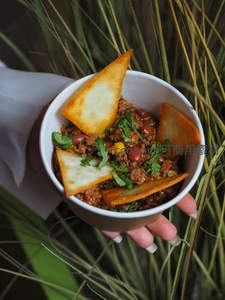 手捧纸杯墨西哥风味肉酱配脆玉米片，背景绿植衬托自然野趣