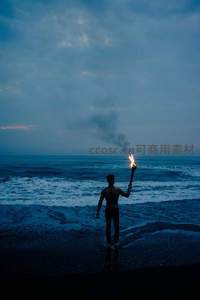 男子海边持火炬伫立，暮色海浪中孤独而坚定的身影