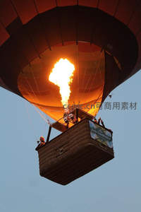 热气球燃烧器喷射烈焰升空，篮中乘客仰望，橙红光晕映染蓝天