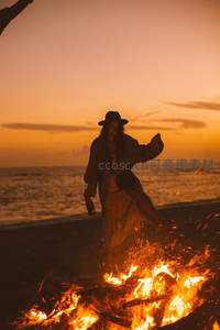 夕阳海滩篝火旁，戴帽女子举瓶起舞剪影