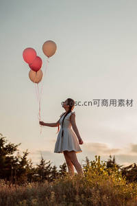 少女手握三只气球仰望天空，金色夕阳洒满田野与裙摆