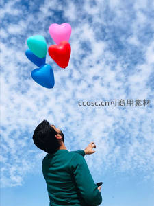 男子仰望五彩心形气球升空，蓝天白云映衬自由瞬间
