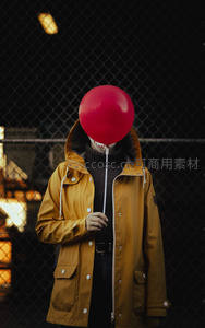 黄雨衣人物持红气球遮面，铁丝网后夜色神秘氛围照