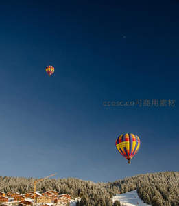 雪山小镇上空双热气球翱翔，彩绘球体映衬松林与木屋冬景