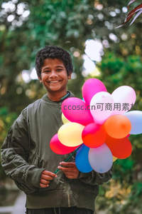 笑容灿烂的男孩手捧彩色气球立于绿荫下