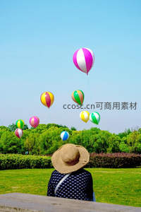 草地上戴草帽的人凝望彩色气球升空，绿树蓝天映衬童趣时光