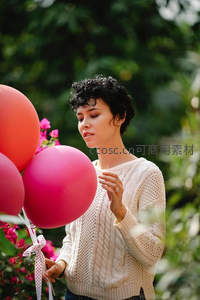 花园中女子轻抚粉色气球，绿植环绕笑容温柔自然