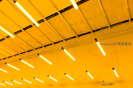 工业风展厅天花板悬挂多组荧光灯管，暖黄基调指引‘GALLERIES’方向