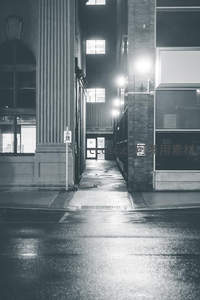雨夜城市窄巷黑白影像，湿漉路面倒映建筑与灯光