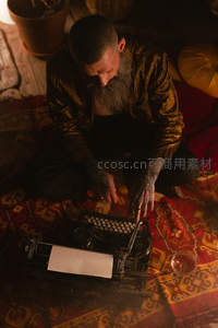 蓄须纹身男子盘坐地毯前专注操作老式打字机，暖光营造神秘氛围